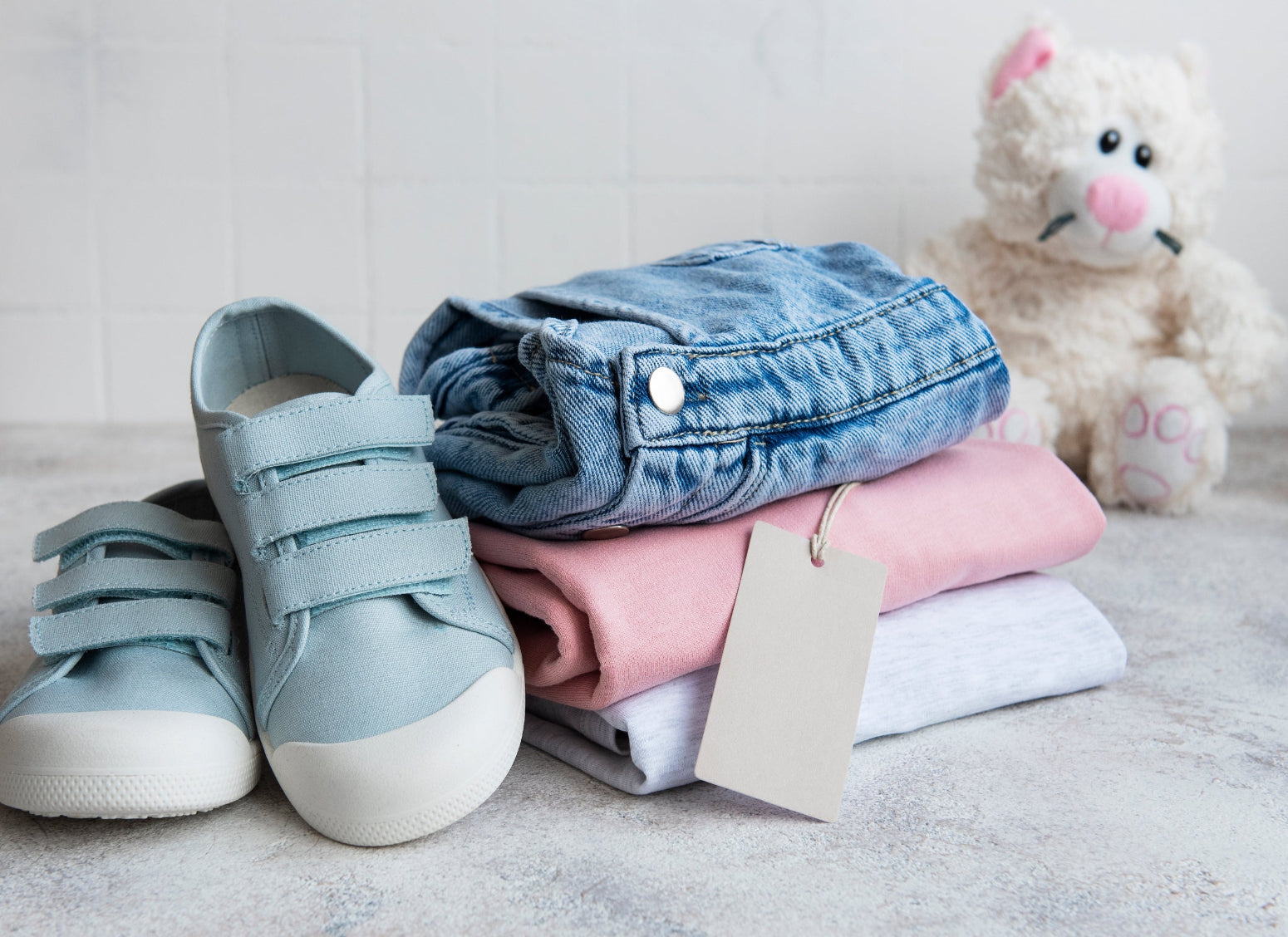 Children's shoes and folded clothes with a tag on a light surface, next to a plush toy.